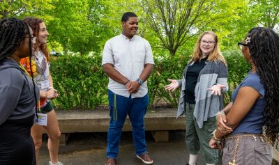 A woman speaks to four students standing in a circle in a wooded seating area.